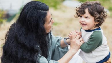 
		Mother crouches in front of her small child and grabs his arms, child looks happily to the side
	